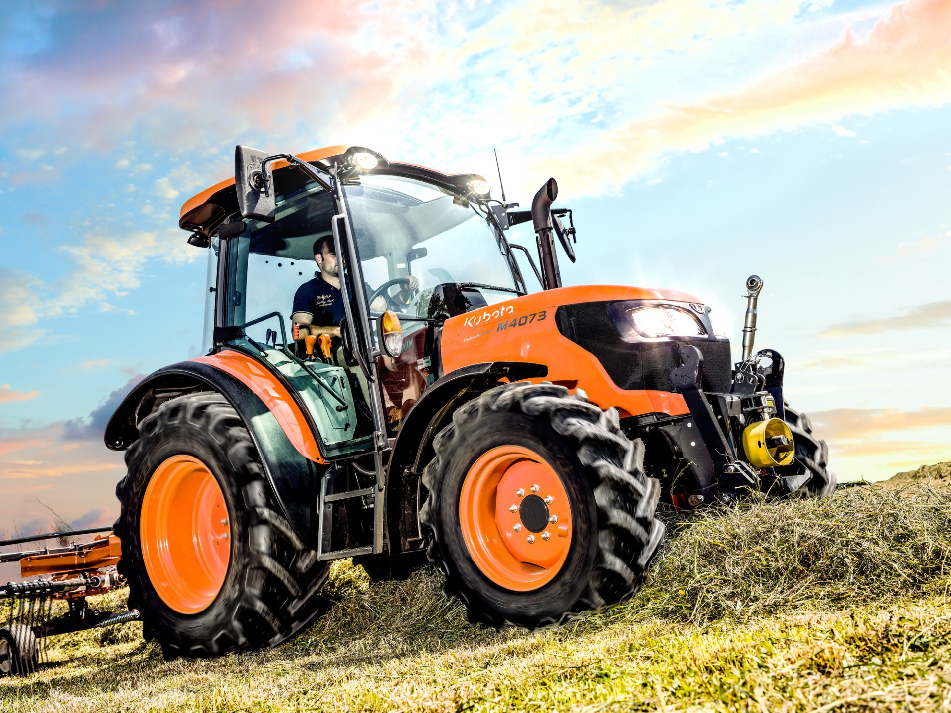Ciągnik Kubota M4-073 ze zgrabiarką na łące, widok z przodu
